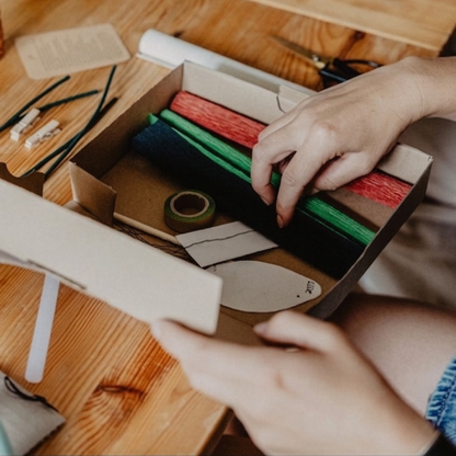 Hände beim Auspacken eines DIY Bastelsets von Woodwormis World mit Krepppapier, Floristendraht und Schablonen auf einem Holztisch